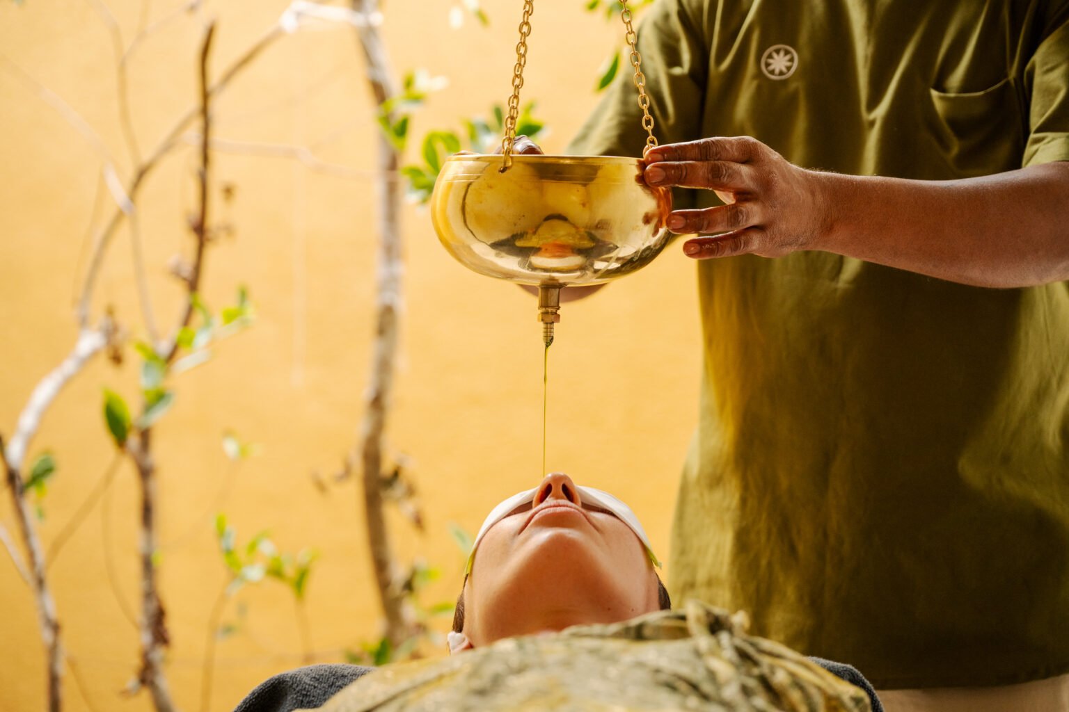 A practitioner pours oil from a brass vessel during a Shirodhara treatment